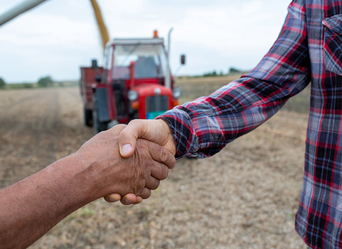 About Our Agency - Close-up of Two People Shaking Hands at a Crop Field With a Red Tractor in the Background